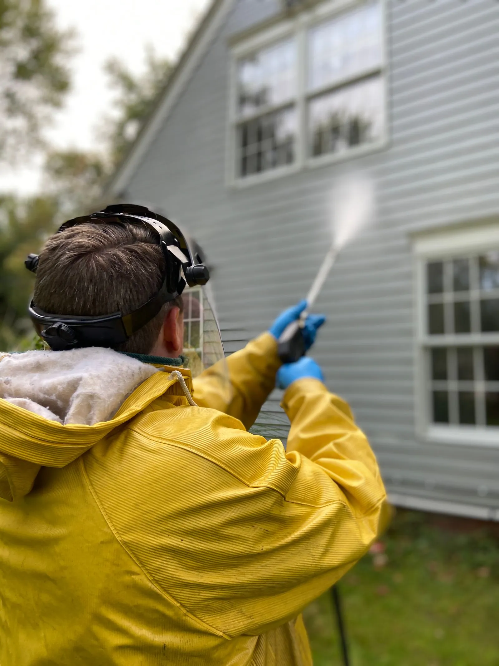 A technician in a rain jacket soft washing the siding of a house
